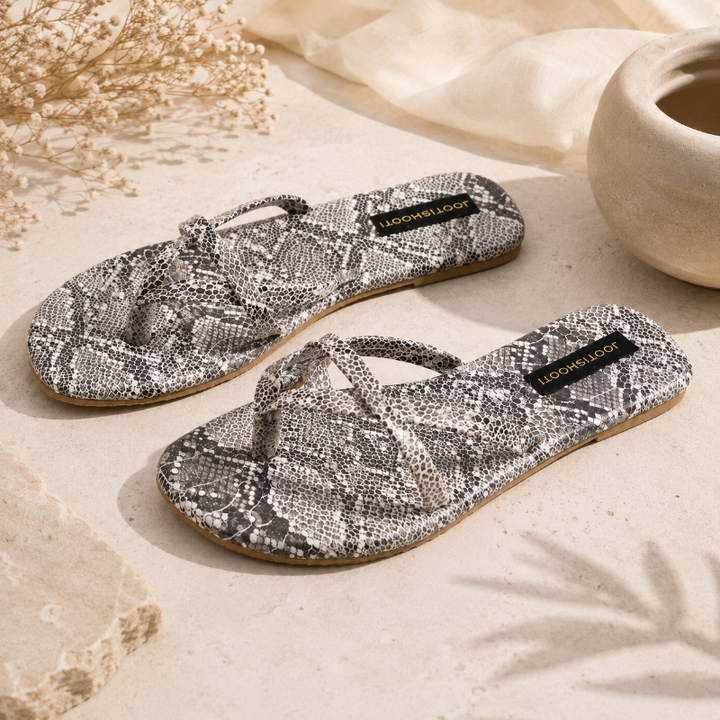 A pair of flat thong sandals with a gray and white snakeskin pattern, displayed on a textured beige surface with dried flowers and a ceramic pot.