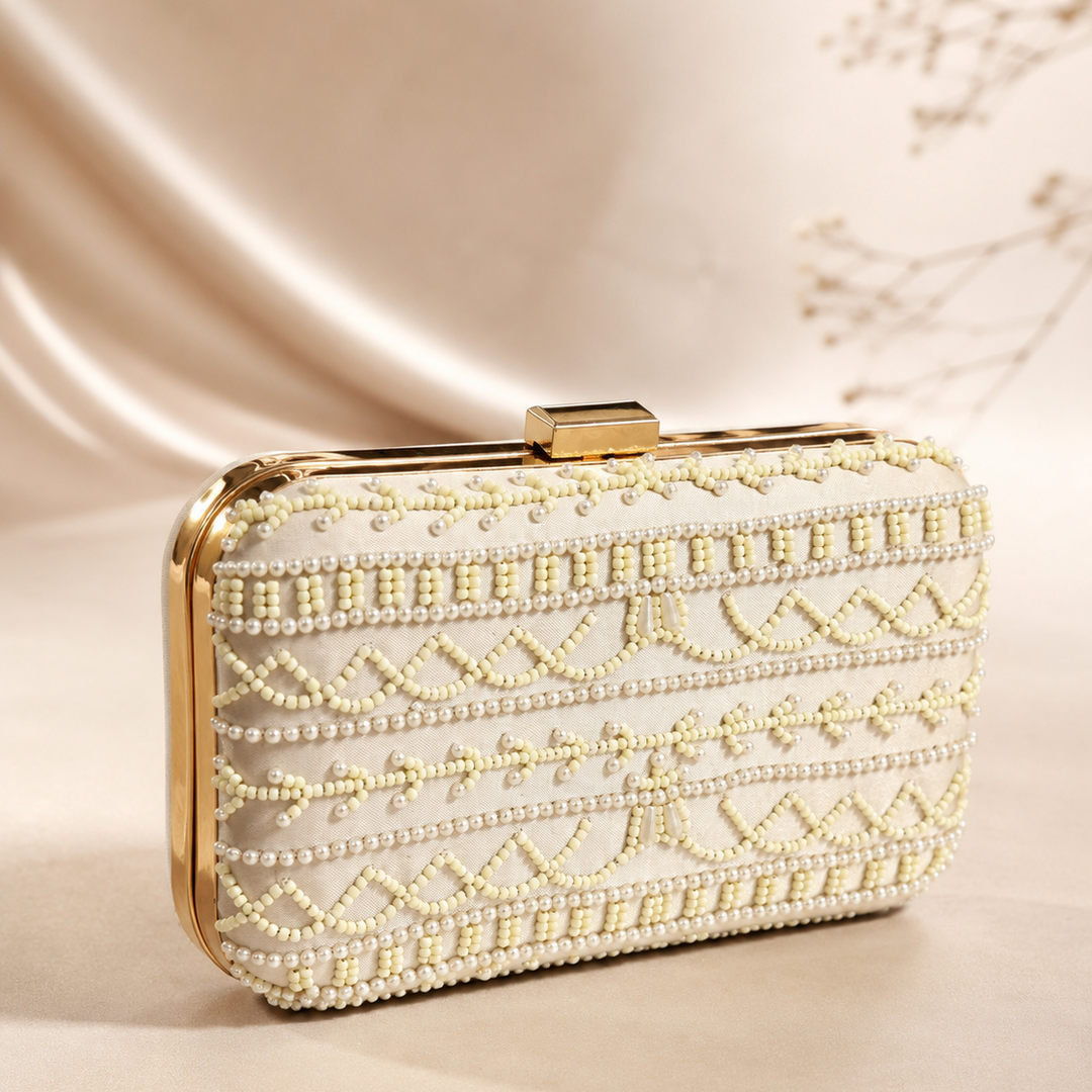 A rectangular clutch bag with a cream-colored faux leather body, adorned with intricate rows of cream and gold beads forming geometric patterns, featuring a gold-tone metal frame and clasp, set against a draped beige satin background.