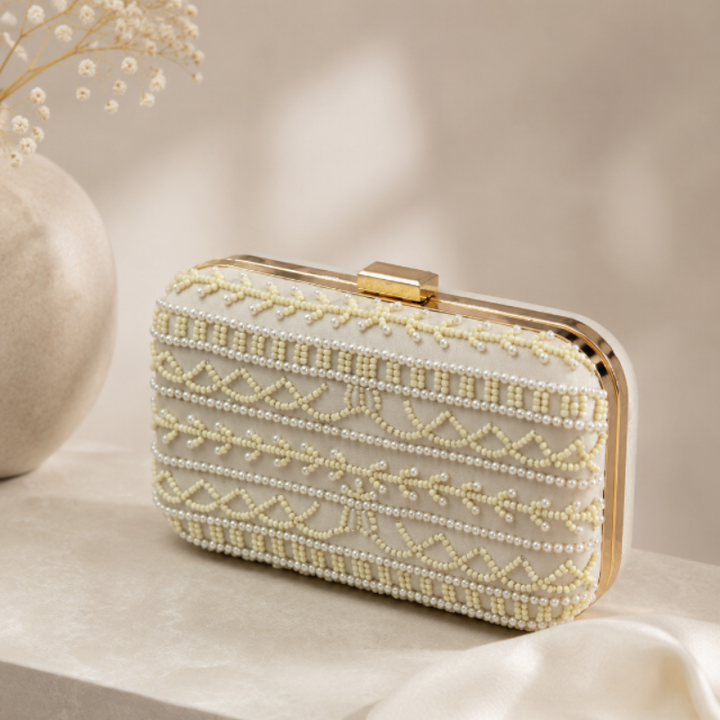 A rectangular evening clutch bag with a cream-colored fabric body, adorned with intricate rows of white and pale yellow beads forming geometric and floral patterns, finished with a gold-tone metal frame and clasp, resting on a light beige stone surface next to a draped white fabric and a decorative vase.