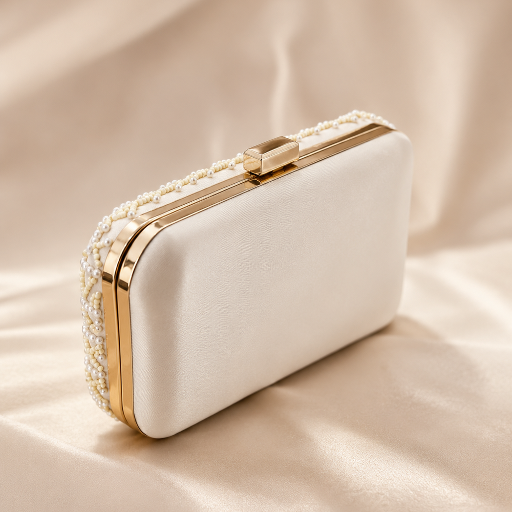 An elegant cream satin evening clutch bag with gold trim and pearl embellishments, resting on a draped beige satin fabric background.