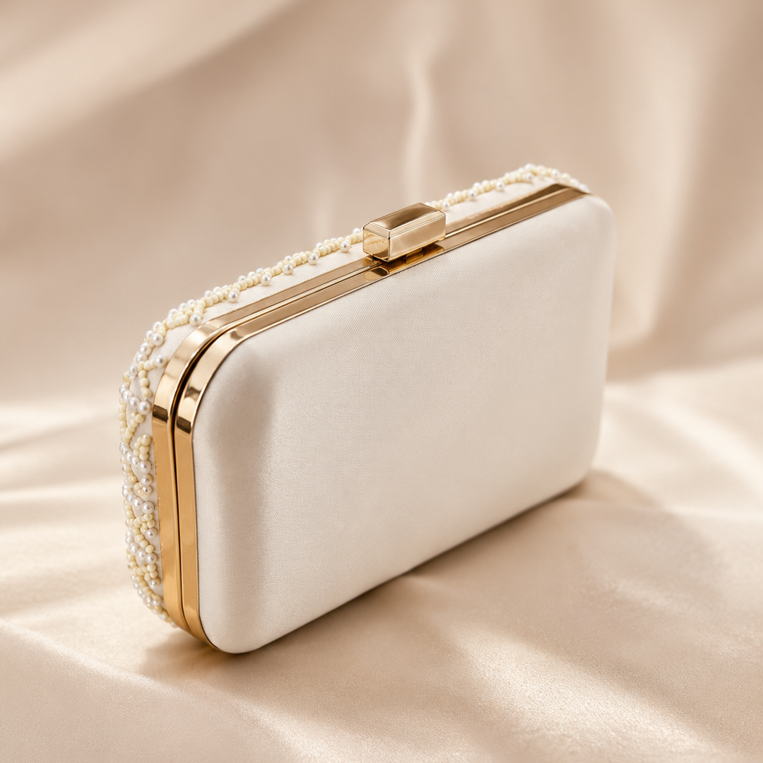 An elegant cream satin evening clutch bag with gold trim and pearl embellishments, resting on a draped beige satin fabric background.