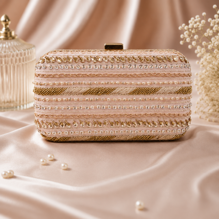 A rectangular evening clutch bag with ornate horizontal stripes of gold and silver beads, sequins, and faux pearls, resting on a draped satin fabric backdrop.