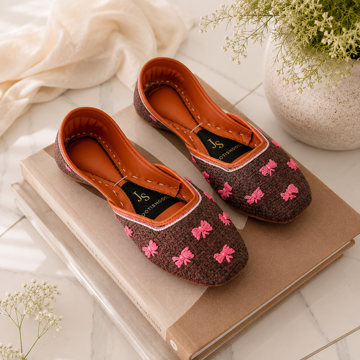 A pair of brown knit khussas with pink bow patterns and orange lining, styled on a stack of books with a neutral fabric and plant in the background.