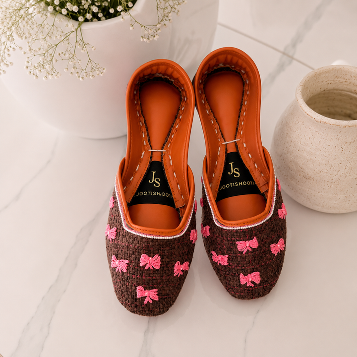 A pair of khussas with brown woven-texture uppers decorated with pink bow motifs, bright orange interiors, and black insoles branded &