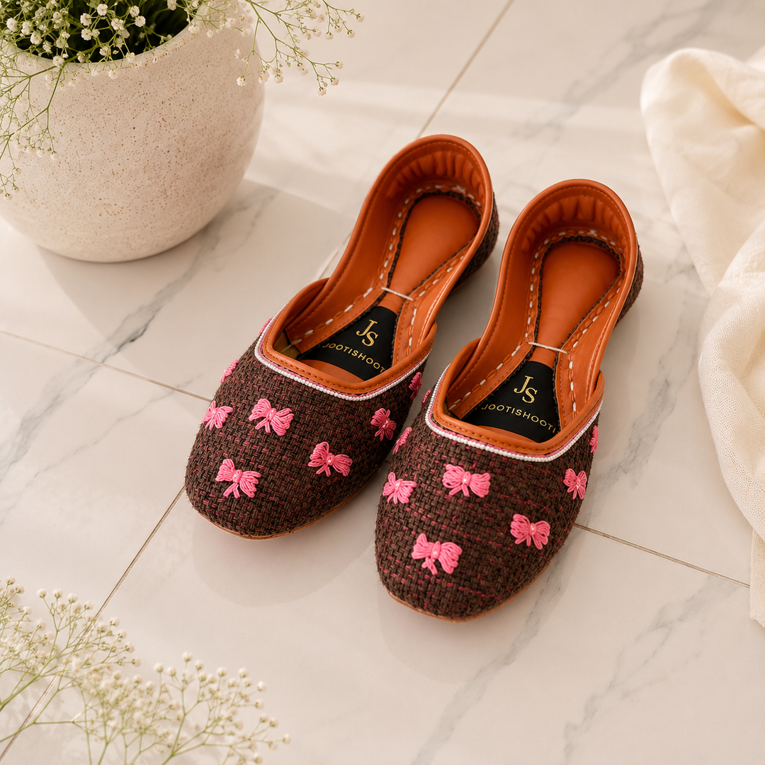A pair of khussas with a brown woven fabric upper, decorated with pink embroidered bows, resting on a light marble tile floor next to a white ceramic planter and a white fabric.
