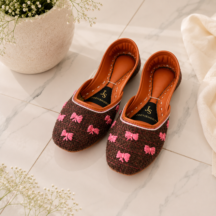 A pair of khussas with a brown woven fabric upper, decorated with pink embroidered bows, resting on a light marble tile floor next to a white ceramic planter and a white fabric.