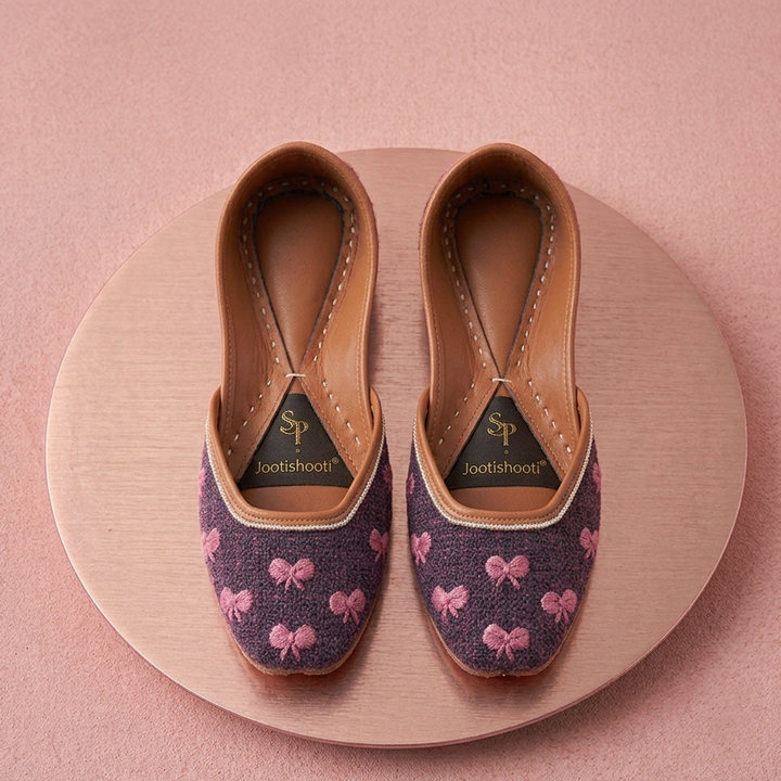 A pair of Jootishooti khussas with a dark blue fabric body decorated with pink bow patterns, displayed on a round wooden platform against a soft pink background.