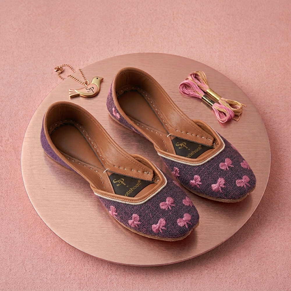 A pair of nay blue khussas with pink bow embroidery, displayed on a round wooden tray with pink thread and a small gold bird charm against a pink background.