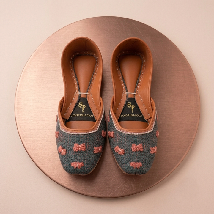 A pair of khussas with a blue-gray woven upper, pink embroidered bow pattern, and tan leather trim, displayed on a round metallic rose-gold platform against a light beige background. The insole shows the brand name &